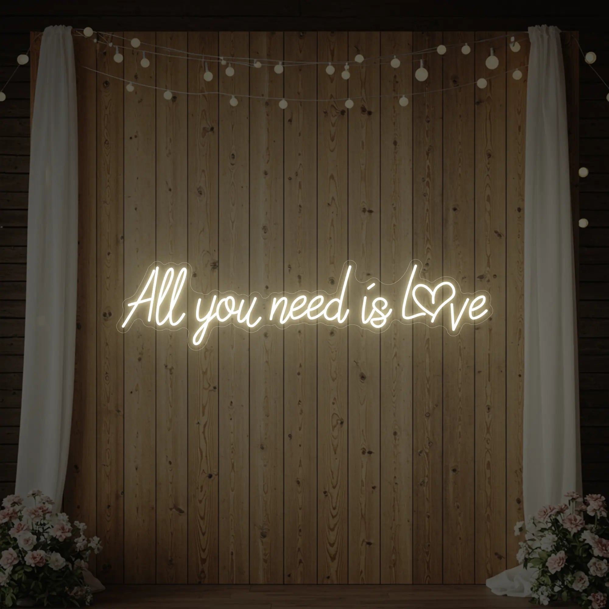 Photo of the 'All You Need Is Love Heart' neon sign glowing in warm white against a styled interior wall.