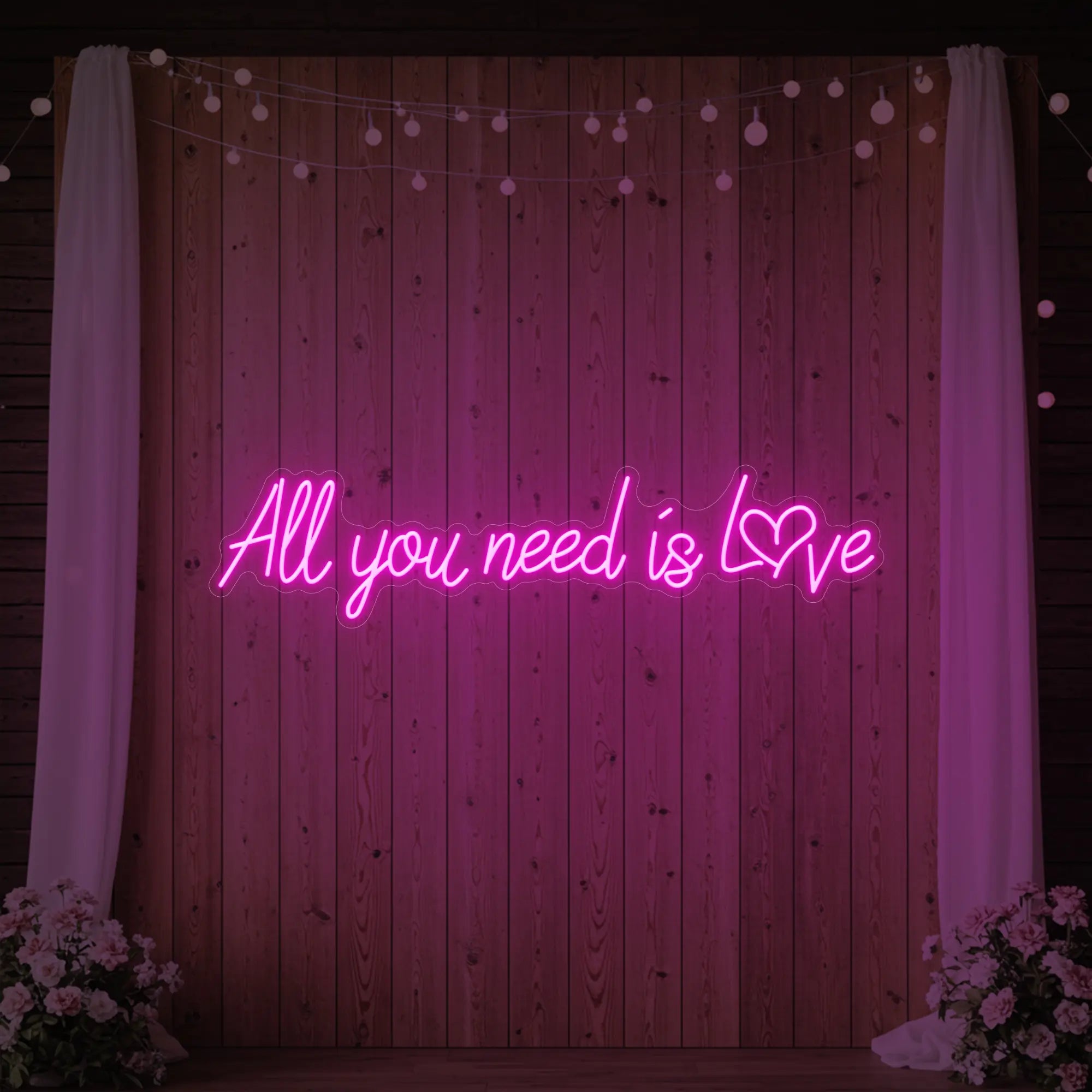 Photo of the 'All You Need Is Love Heart' neon sign glowing in light pink against a styled interior wall.