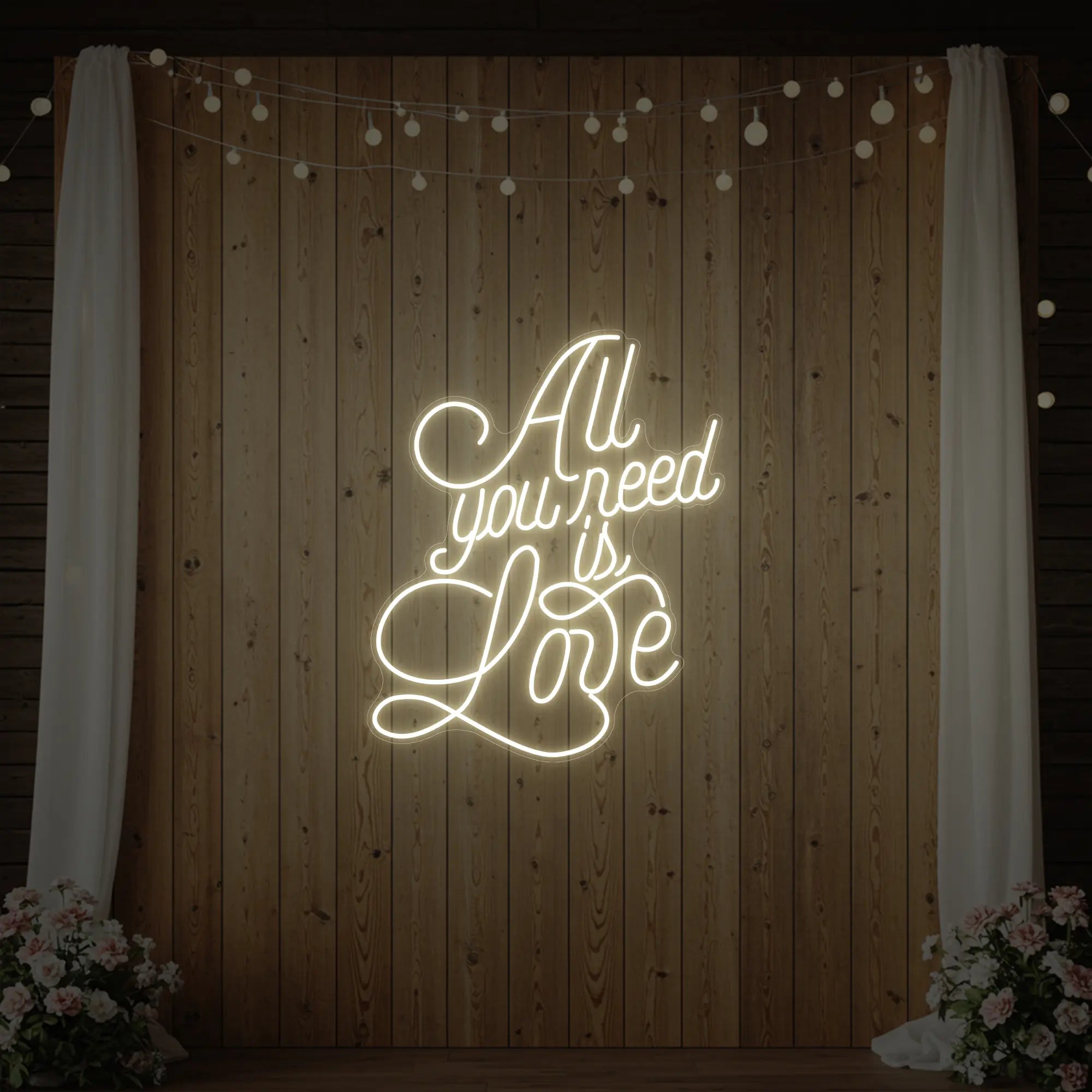 Photo of the 'All You Need Is Love V2' neon sign glowing in warm white against a styled interior wall.