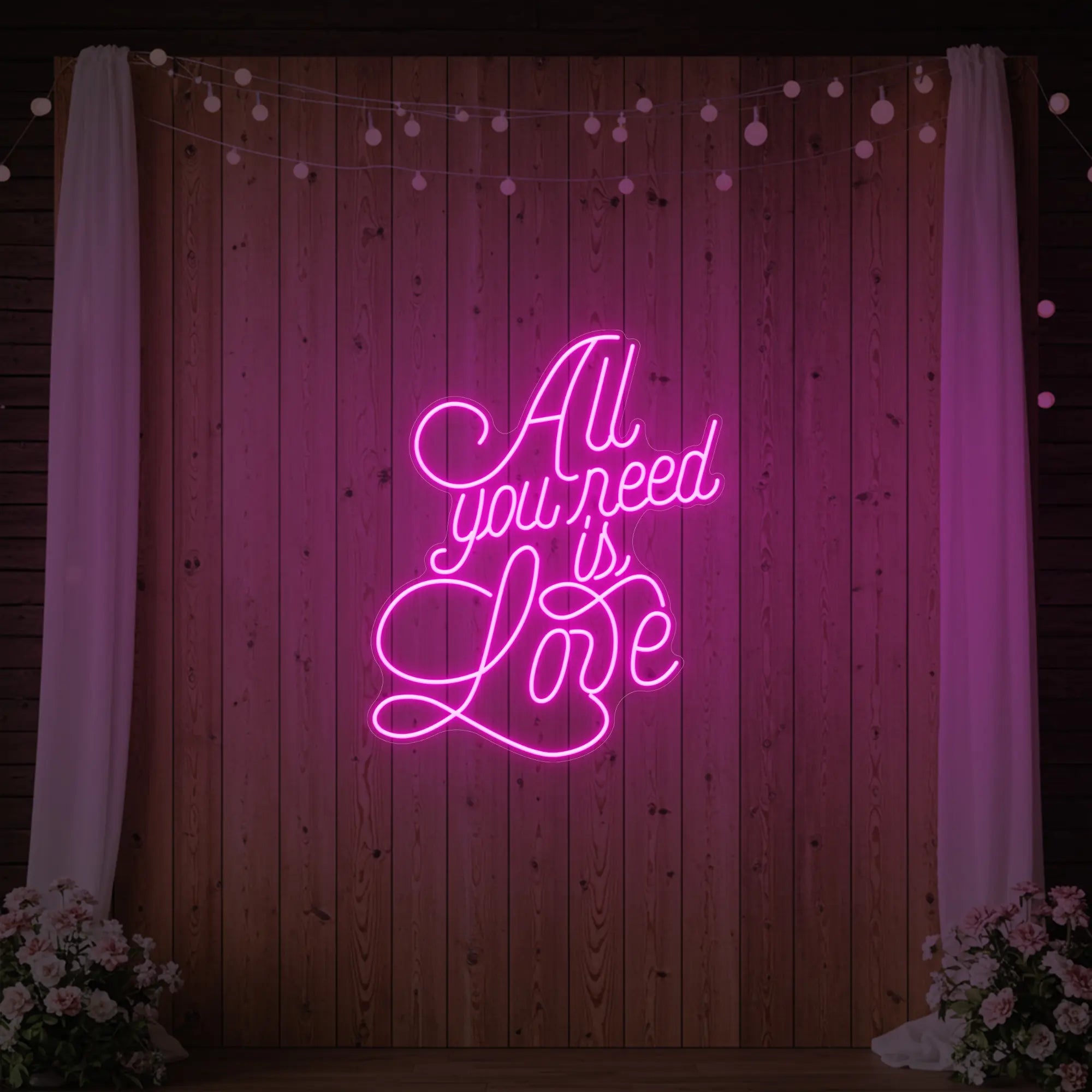 Photo of the 'All You Need Is Love V2' neon sign glowing in light pink against a styled interior wall.
