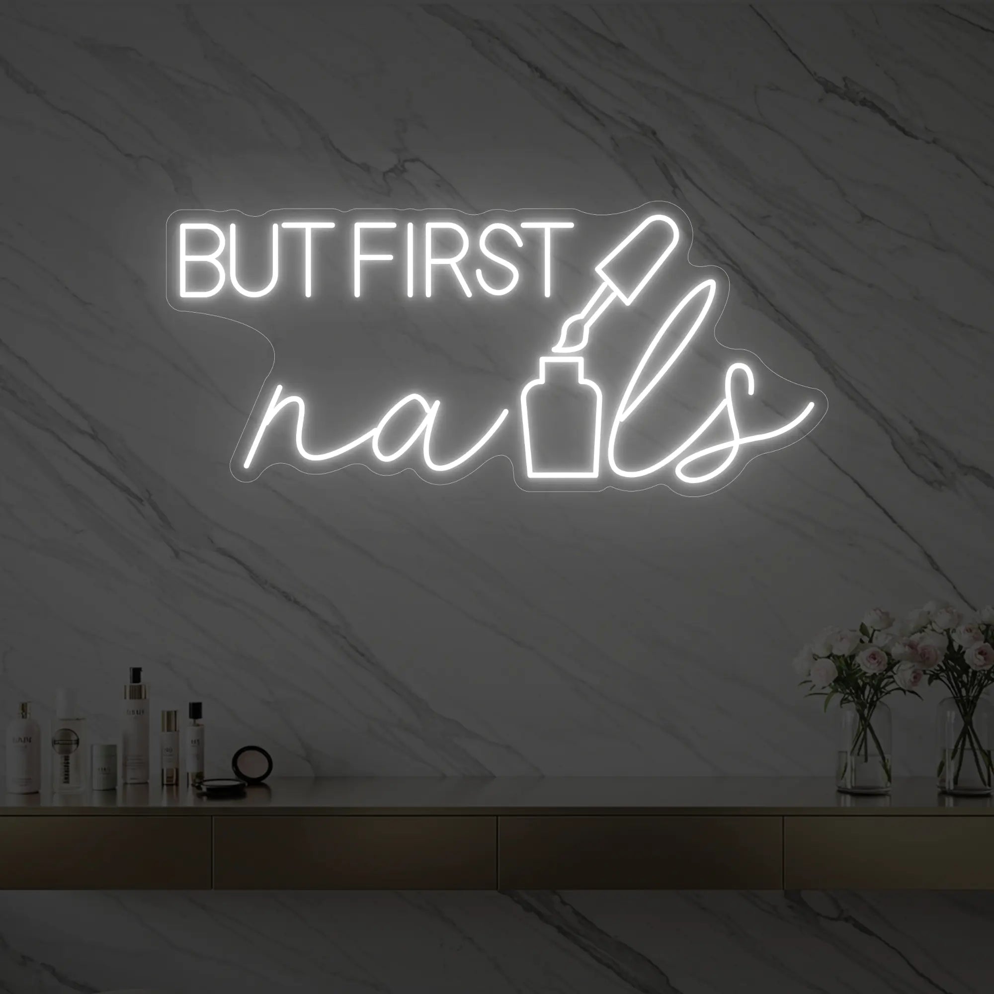 Photo of the 'BUT FIRST nails' neon sign with a nail polish bottle graphic glowing in cool white against a styled interior wall.