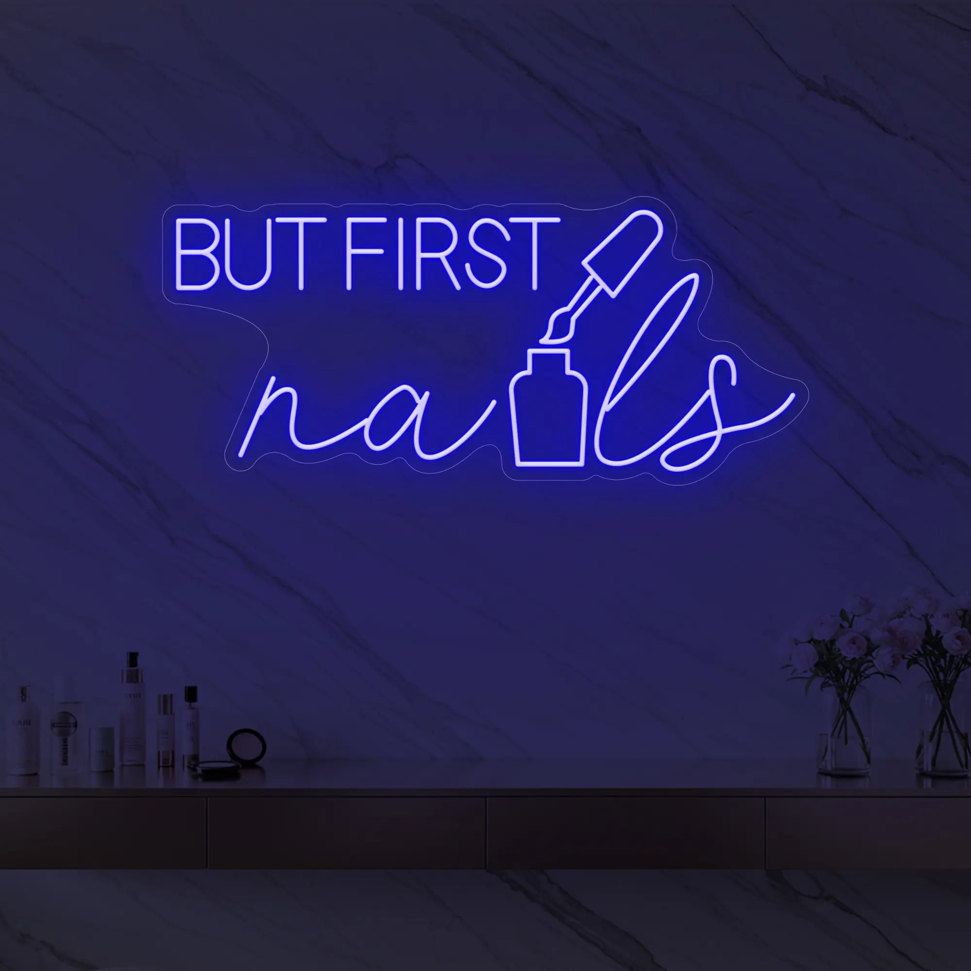 Photo of the 'BUT FIRST nails' neon sign with a nail polish bottle graphic glowing in deep blue against a styled interior wall.