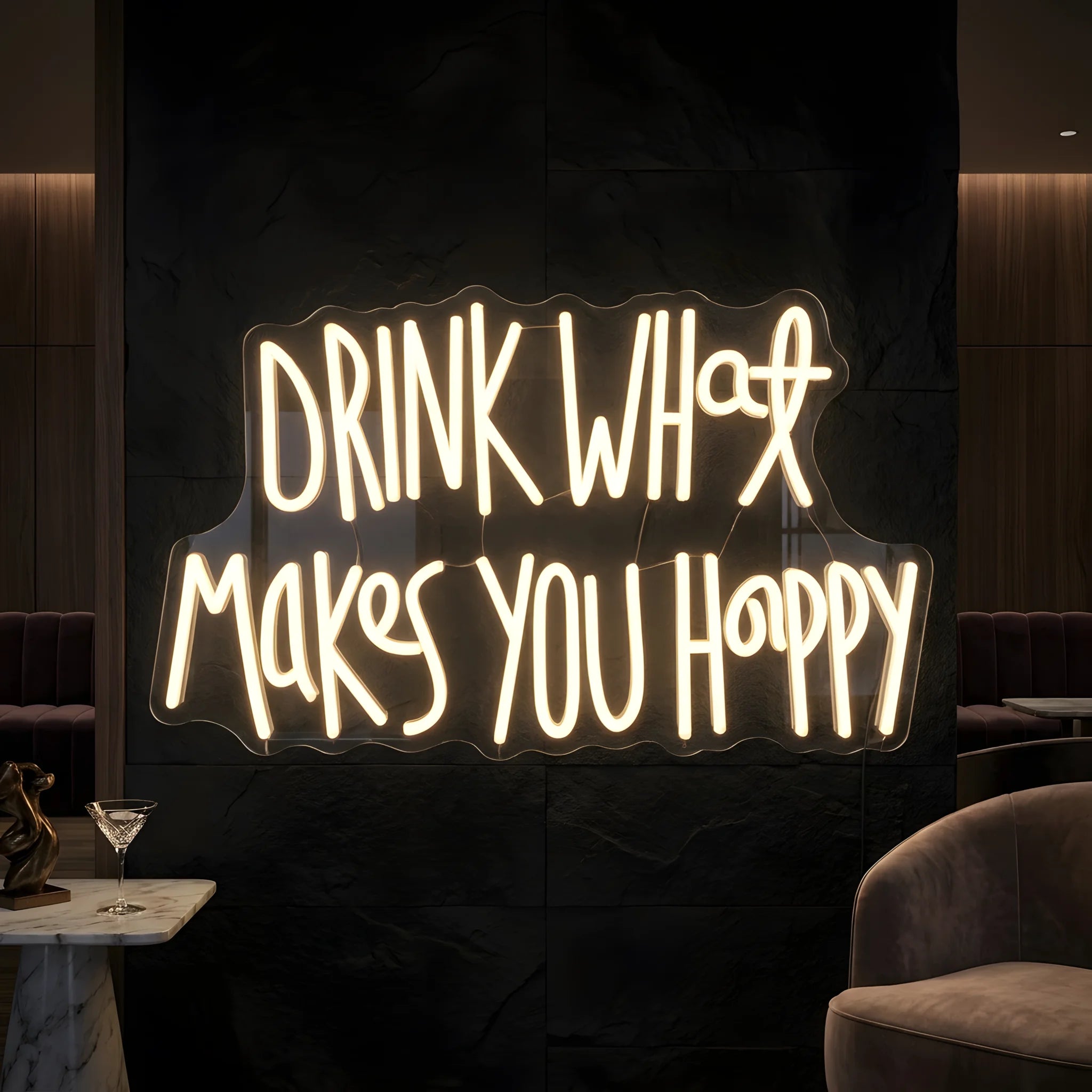 Photo of the 'Drink What Makes You Happy' neon sign glowing in warm white against a styled interior wall.