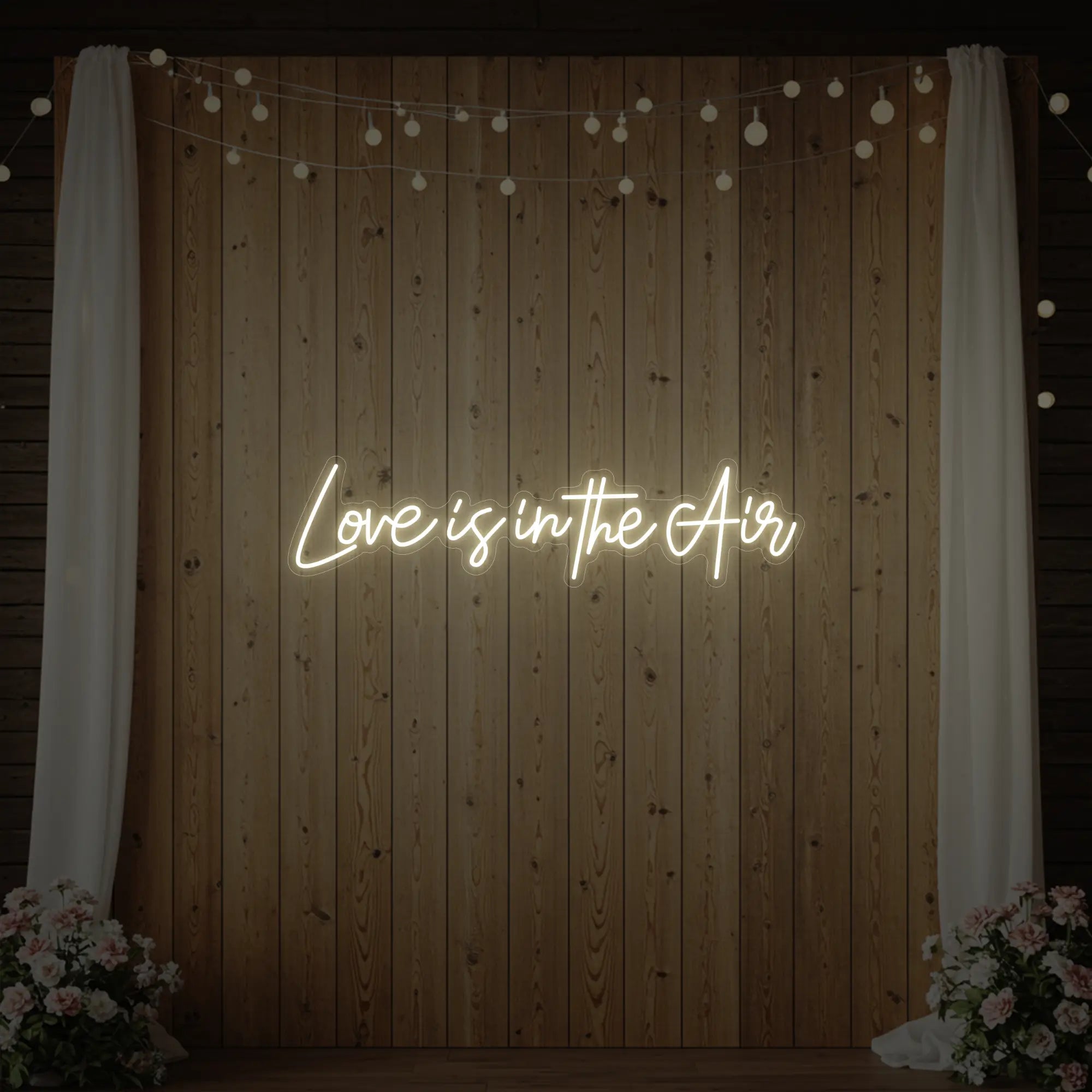 Photo of the 'Love Is In The Air' neon sign glowing in warm white against a styled interior wall.