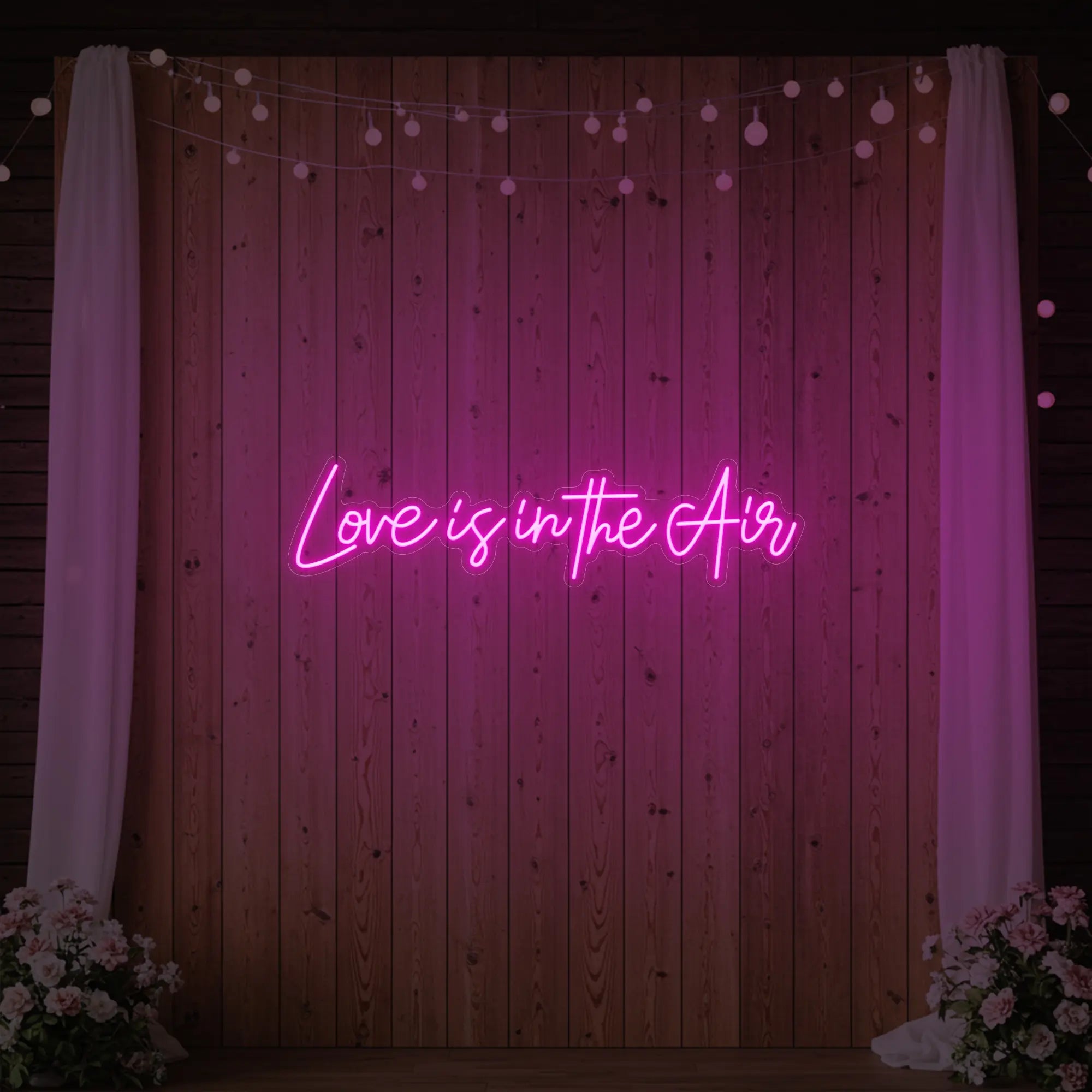 Photo of the 'Love Is In The Air' neon sign glowing in light pink against a styled interior wall.