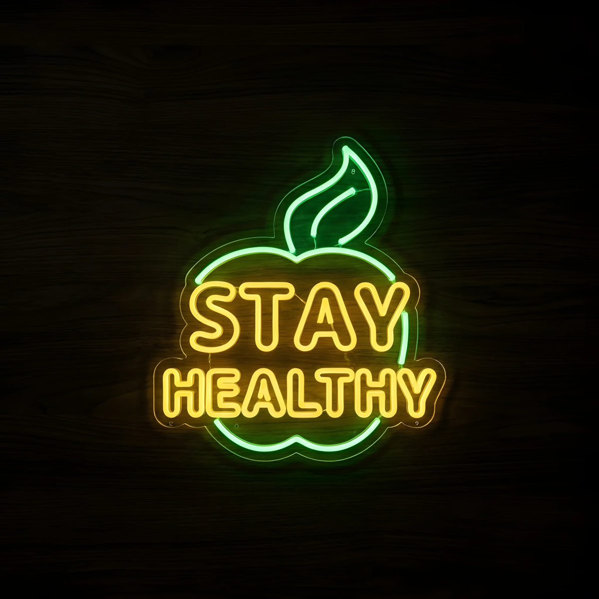 Stay Healthy Leaf LED Neon Sign featuring bright yellow STAY HEALTHY lettering inside a glowing green apple outline with leaf on clear acrylic backing against a dark wooden wall.
