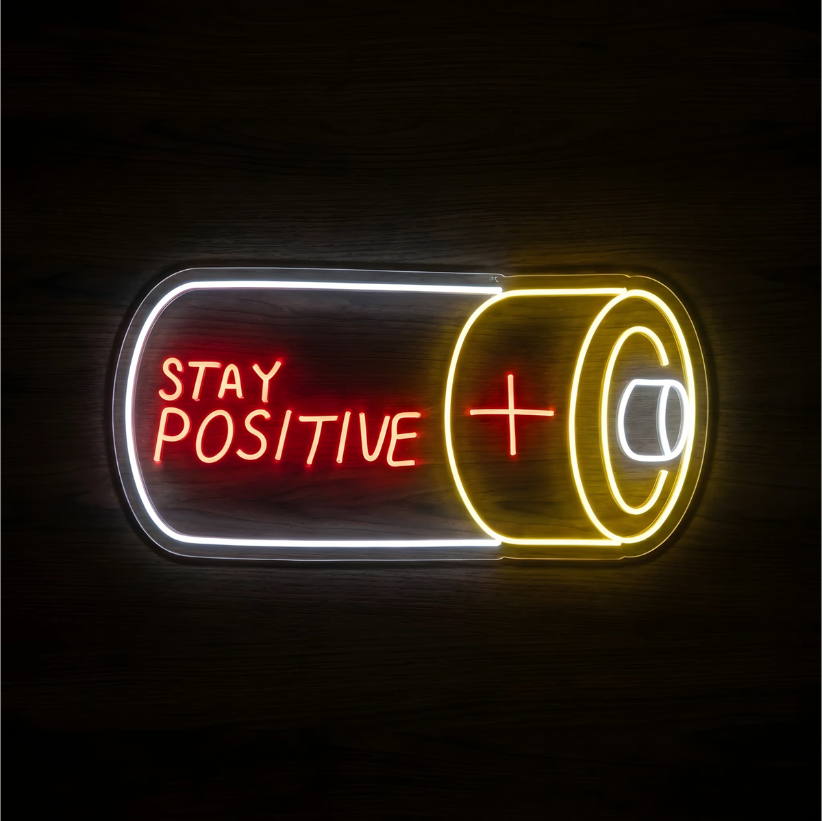 Stay Positive LED Neon Sign with red lettering, a yellow plus sign, a smiling face, and a white outline in a capsule shape on clear acrylic against a dark wood background, suited to bedrooms and home offices.