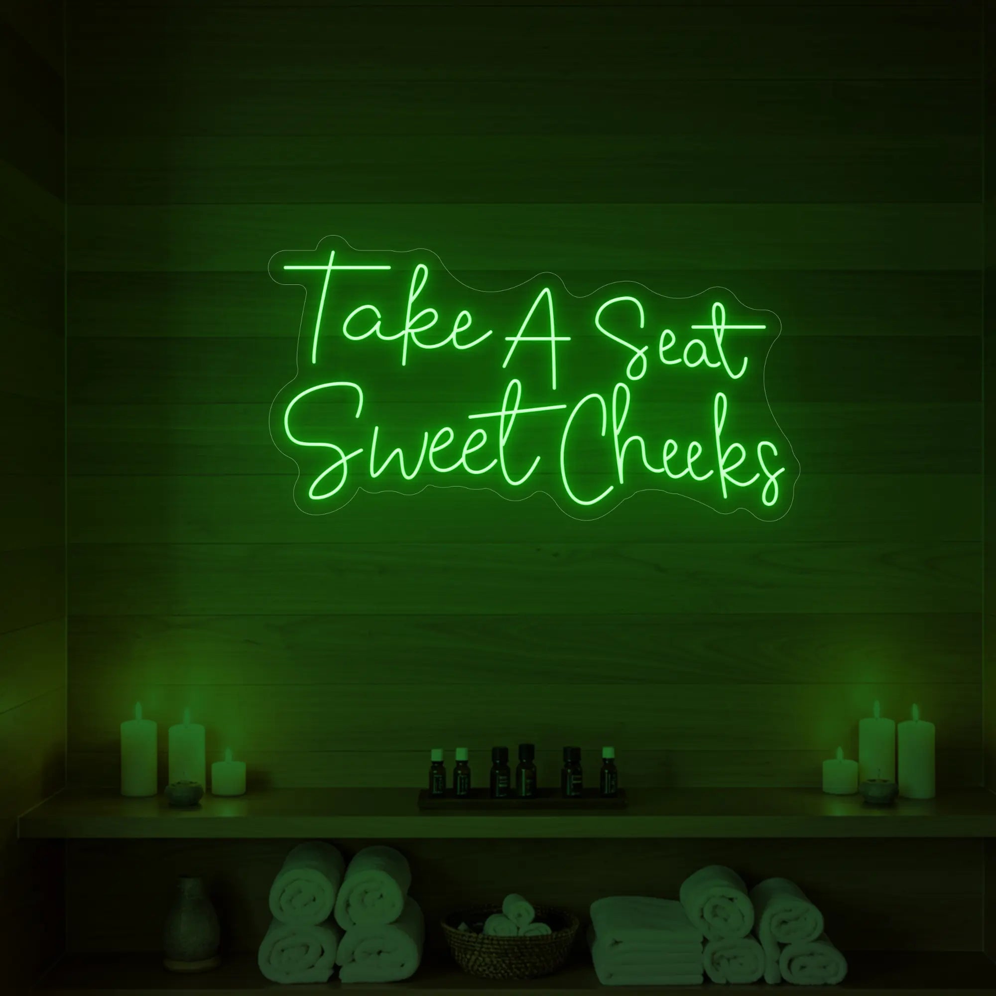 Photo of the 'Take A Seat Sweet Cheeks' neon sign glowing in green against a styled interior wall.