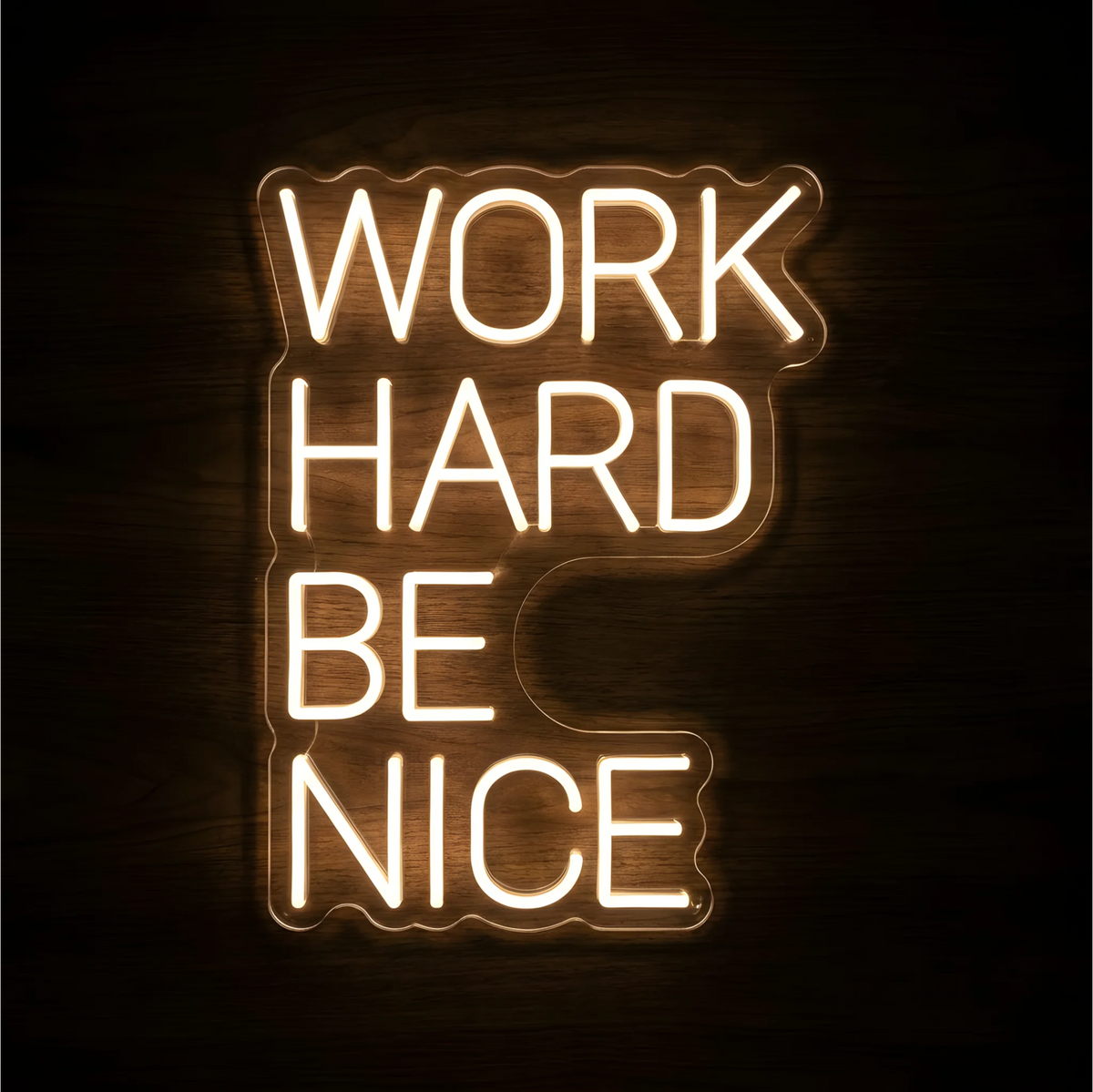 “WORK HARD BE NICE” LED Neon Sign glowing in warm white lettering on clear acrylic backing against a dark wood background, suitable for office, classroom, and reception area decor.