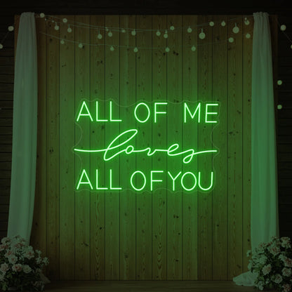 Photo of the 'All Of Me Love All Of You' neon sign glowing in green against a styled interior wall.