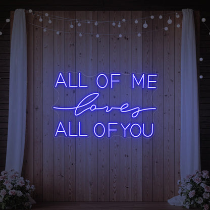 Photo of the 'All Of Me Love All Of You' neon sign glowing in deep blue against a styled interior wall.