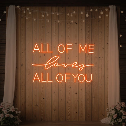 Photo of the 'All Of Me Love All Of You' neon sign glowing in golden yellow against a styled interior wall.