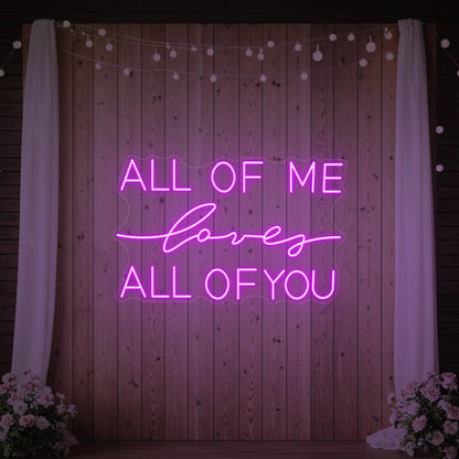 Photo of the 'All Of Me Love All Of You' neon sign glowing in deep pink against a styled interior wall.