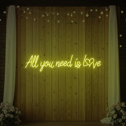 Photo of the 'All You Need Is Love Heart' neon sign glowing in lemon yellow against a styled interior wall.