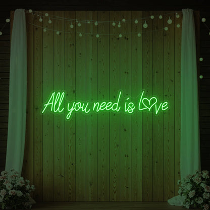 Photo of the 'All You Need Is Love Heart' neon sign glowing in green against a styled interior wall.