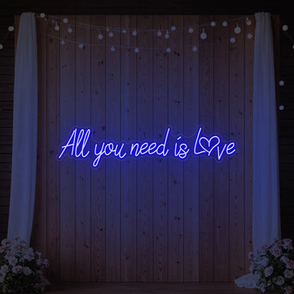Photo of the 'All You Need Is Love Heart' neon sign glowing in deep blue against a styled interior wall.