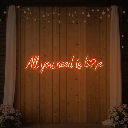 Photo of the 'All You Need Is Love Heart' neon sign glowing in golden yellow against a styled interior wall.