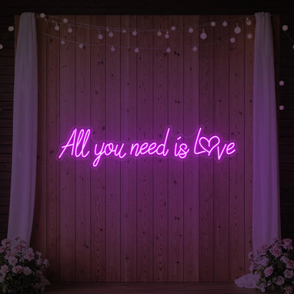 Photo of the 'All You Need Is Love Heart' neon sign glowing in deep pink against a styled interior wall.