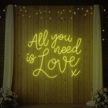 Photo of the 'All You Need Is Love V1' neon sign glowing in lemon yellow against a styled interior wall.
