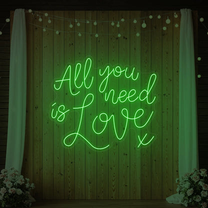 Photo of the 'All You Need Is Love V1' neon sign glowing in green against a styled interior wall.