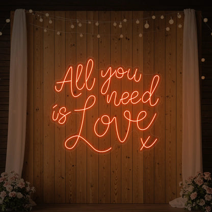 Photo of the 'All You Need Is Love V1' neon sign glowing in golden yellow against a styled interior wall.