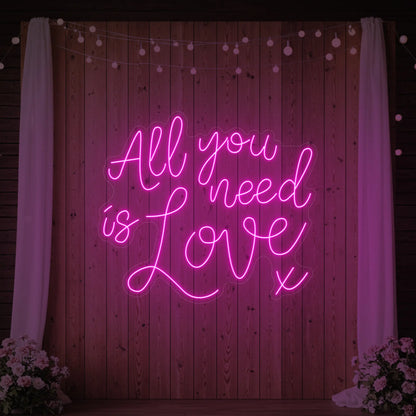 Photo of the 'All You Need Is Love V1' neon sign glowing in light pink against a styled interior wall.