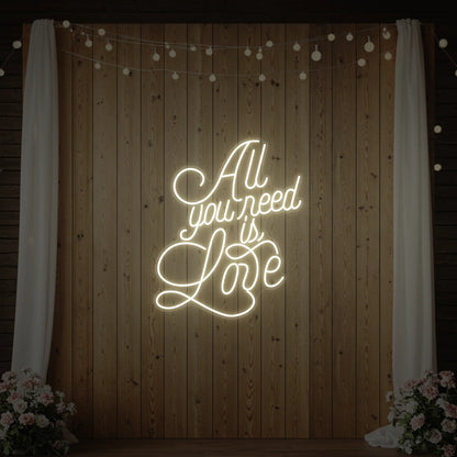 Photo of the 'All You Need Is Love V2' neon sign glowing in warm white against a styled interior wall.