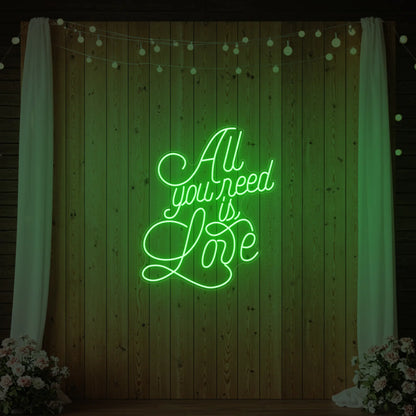 Photo of the 'All You Need Is Love V2' neon sign glowing in green against a styled interior wall.