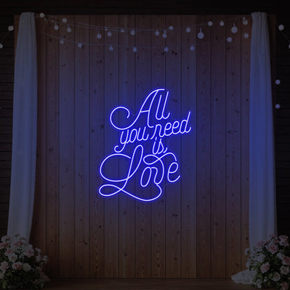 Photo of the 'All You Need Is Love V2' neon sign glowing in deep blue against a styled interior wall.