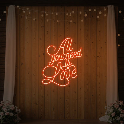 Photo of the 'All You Need Is Love V2' neon sign glowing in golden yellow against a styled interior wall.