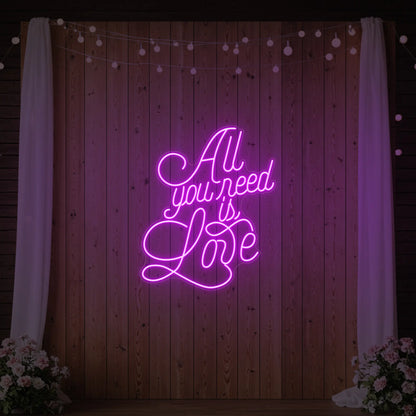 Photo of the 'All You Need Is Love V2' neon sign glowing in deep pink against a styled interior wall.