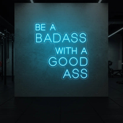  'Be a Badass With a Good Ass' neon sign glowing in ice blue against a styled interior wall.