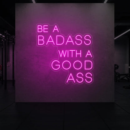  'Be a Badass With a Good Ass' neon sign glowing in light pink against a styled interior wall.