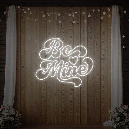 Photo of the 'Be Mine' neon sign glowing in cool white against a styled interior wall.