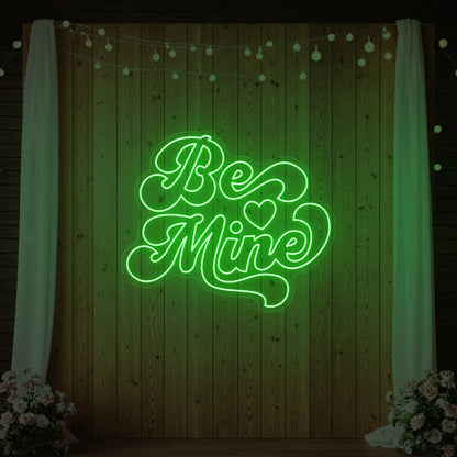 Photo of the 'Be Mine' neon sign glowing in green against a styled interior wall.