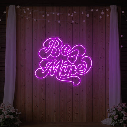 Photo of the 'Be Mine' neon sign glowing in deep pink against a styled interior wall.