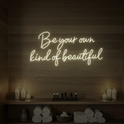 Photo of the 'Be Your Own Kind of Beautiful' neon sign glowing in warm white against a styled interior wall.