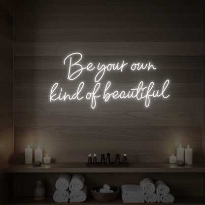 Photo of the 'Be Your Own Kind of Beautiful' neon sign glowing in cool white against a styled interior wall.