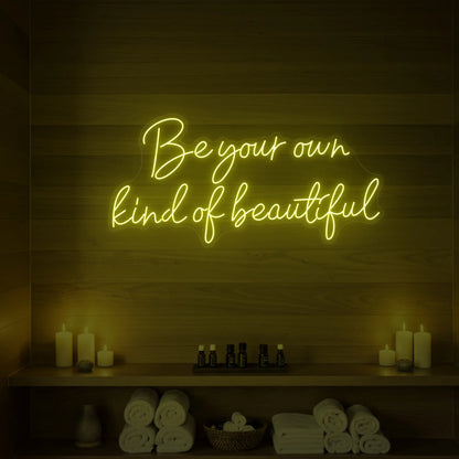 Photo of the 'Be Your Own Kind of Beautiful' neon sign glowing in lemon yellow against a styled interior wall.