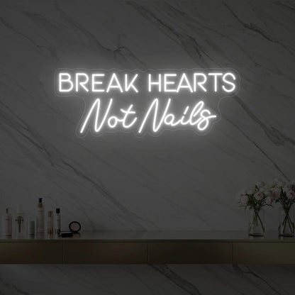 Photo of the 'BREAK HEARTS Not Nails' neon sign glowing in cool white against a styled interior wall.
