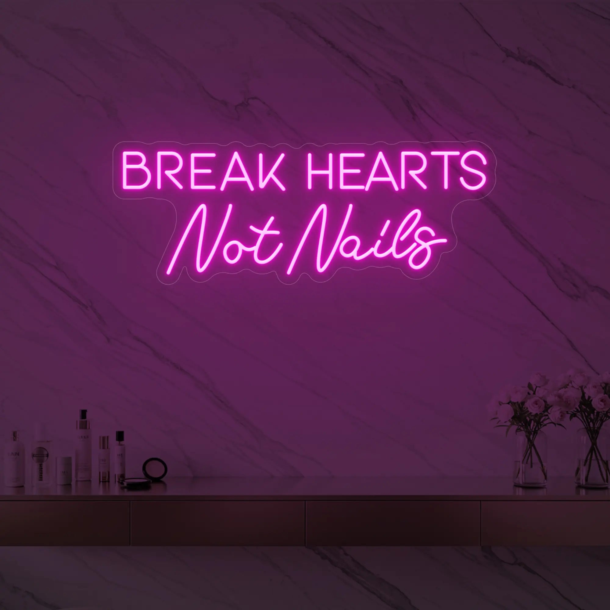 Photo of the 'BREAK HEARTS Not Nails' neon sign glowing in light pink against a styled interior wall.