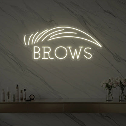 Photo of the 'BROWS' neon sign with an eyebrow graphic glowing in warm white against a styled interior wall.