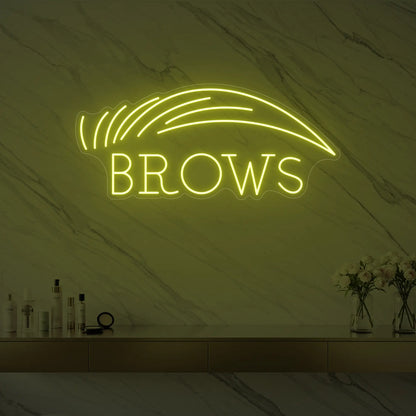 Photo of the 'BROWS' neon sign with an eyebrow graphic glowing in lemon yellow against a styled interior wall.