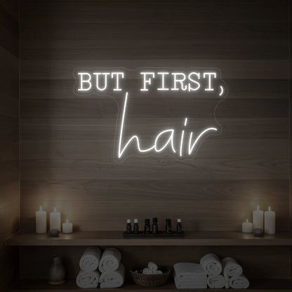 Photo of the 'BUT FIRST, hair' neon sign glowing in cool white against a styled interior wall.