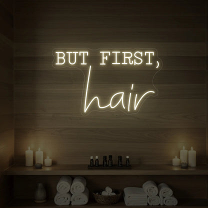 Photo of the 'BUT FIRST, hair' neon sign glowing in warm white against a styled interior wall.