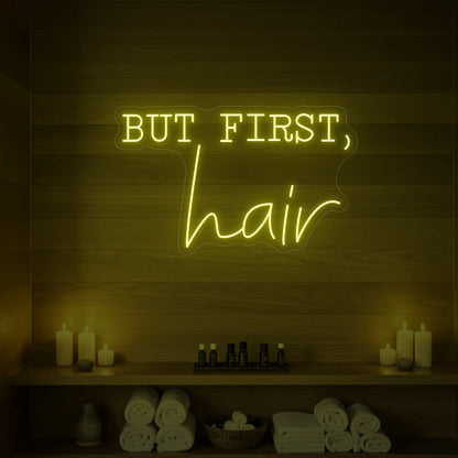 Photo of the 'BUT FIRST, hair' neon sign glowing in lemon yellow against a styled interior wall.