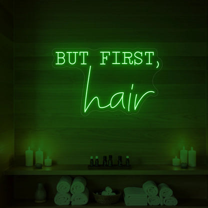 Photo of the 'BUT FIRST, hair' neon sign glowing in green against a styled interior wall.