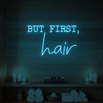 Photo of the 'BUT FIRST, hair' neon sign glowing in ice blue against a styled interior wall.