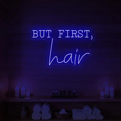 Photo of the 'BUT FIRST, hair' neon sign glowing in deep blue against a styled interior wall.