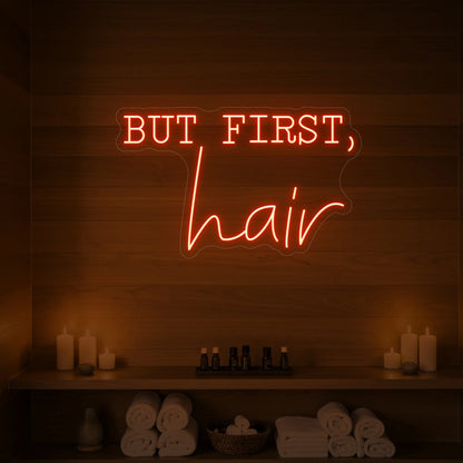 Photo of the 'BUT FIRST, hair' neon sign glowing in golden yellow against a styled interior wall.